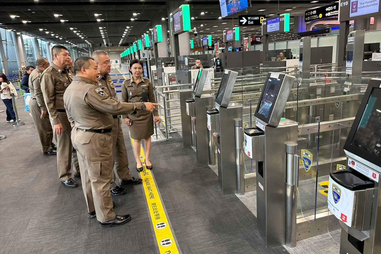 Royal Thai Immigration Bureau officers inspecting automated passport e-gates in an airport arrivals hall.