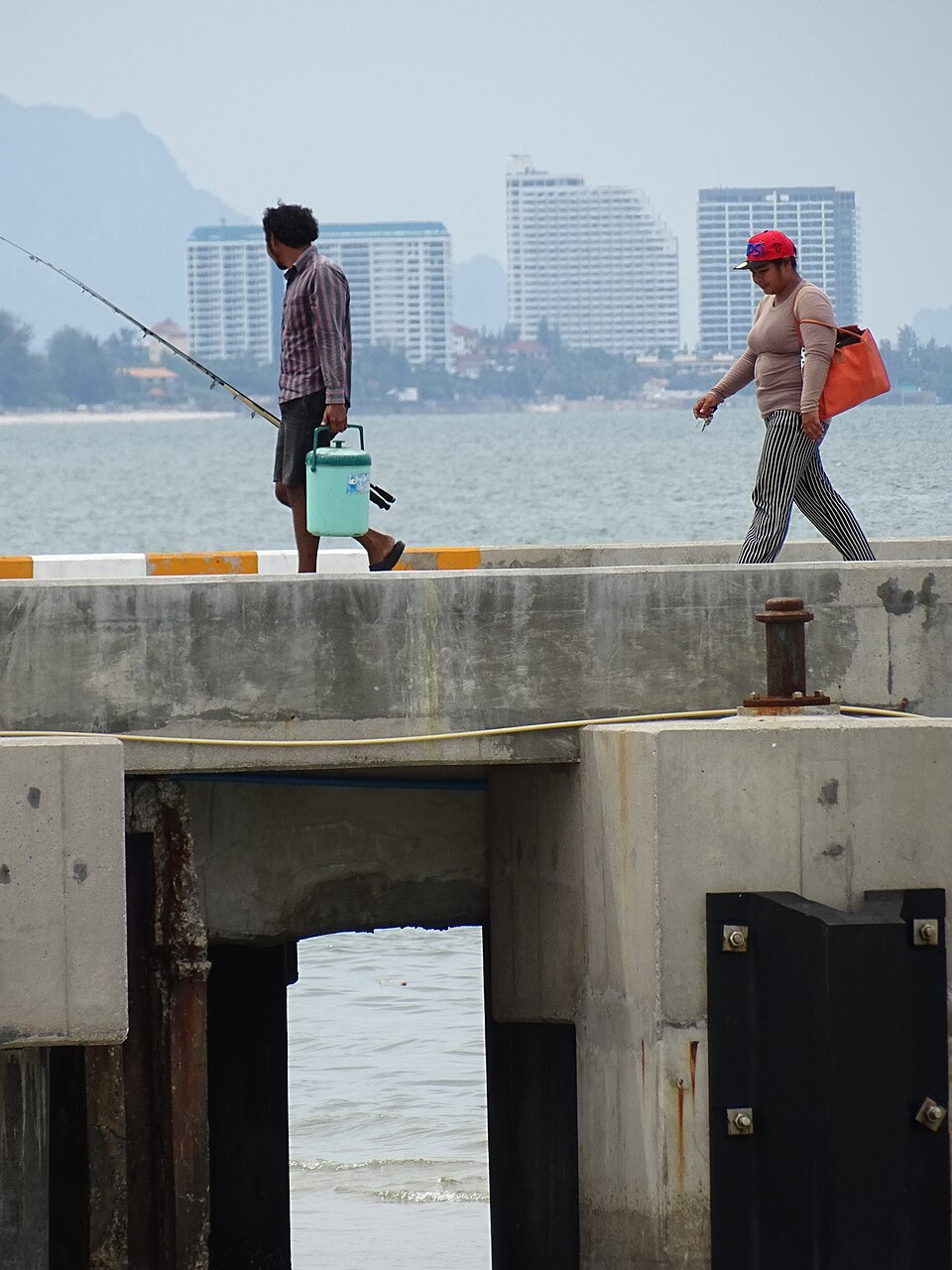 Anglers walking along a concrete pier in Hua Hin, Thailand, with beachfront condo towers in the hazy distance, a view for expats living in Thailand and renting in Hua Hin